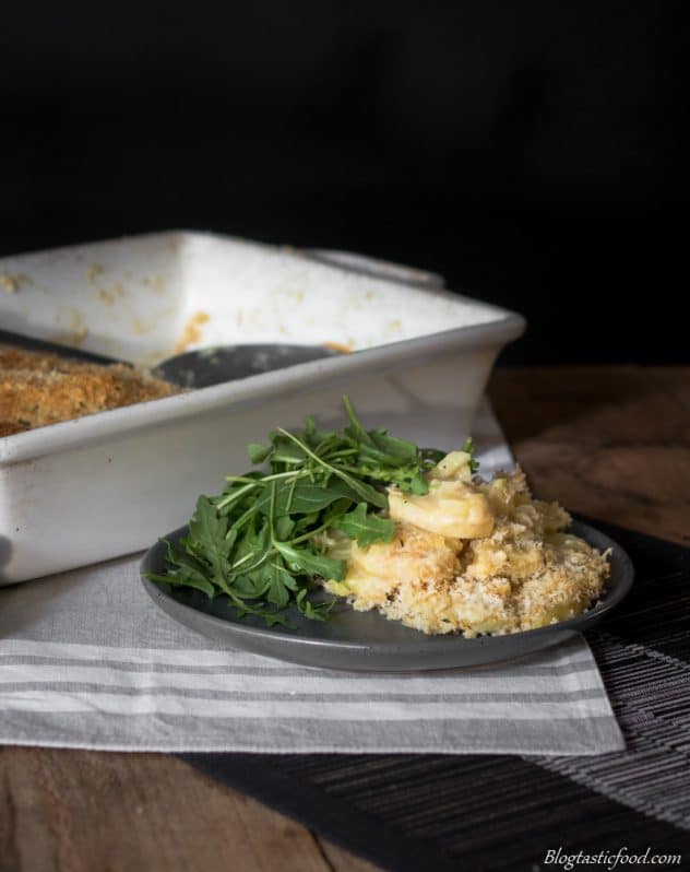 Vegan potato served on a black plate with a side of salad leaves. There is also a white baking tray filled with the vegan potato gratin in the background.