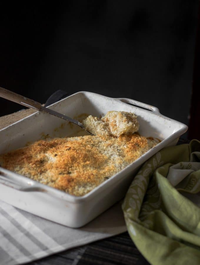 A photo of vegan potato gratin with panko breadcrumb topping served in a baking tray with a large spoon scooping out some of the potato.