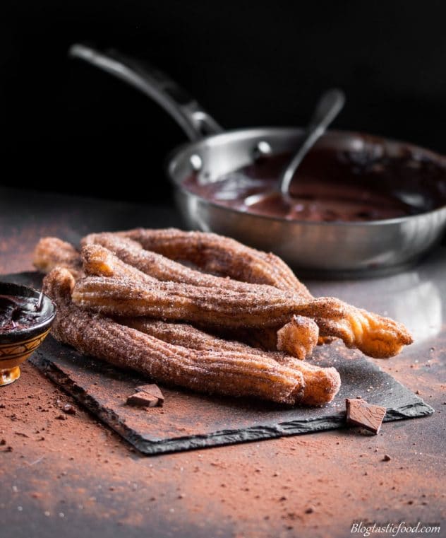 Churros served on a dark sushi platter with a pan of chocolate sauce in the background.