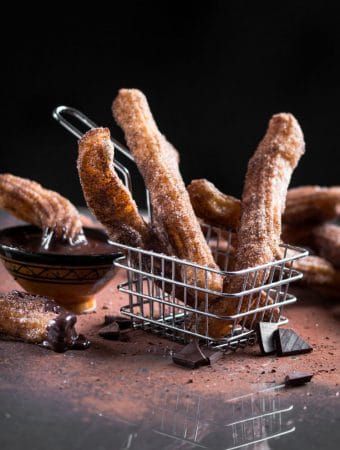 A dark, contrast photo of churros served in a small metal chip basket with cocoa dusted around.
