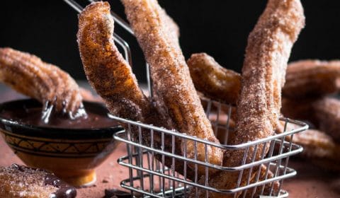 A dark, contrast photo of churros served in a small metal chip basket with cocoa dusted around.