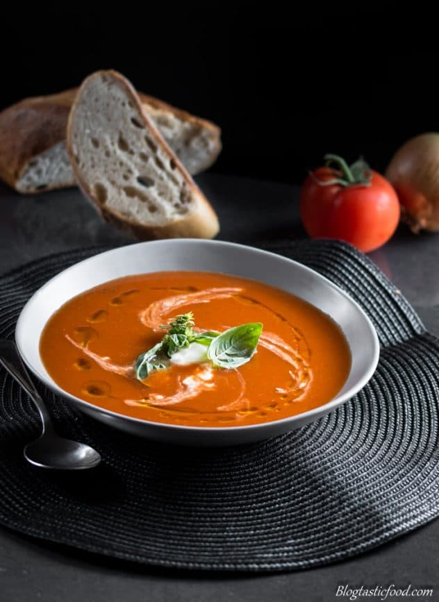 A bowl of spicy tomato soup with bread, an onion and a fresh tomato in the background. 