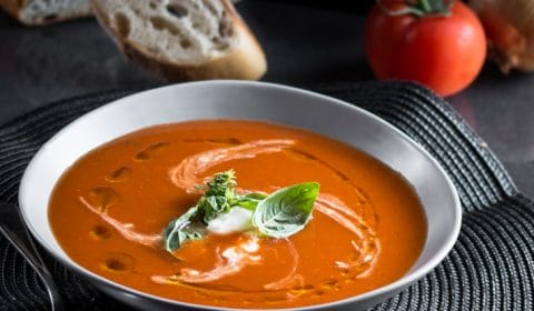 a dark moody photo of a bowl of spicy tomato soup garnished with sour cream and fresh basil leaves.