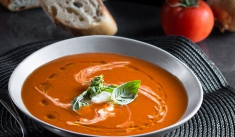 a dark moody photo of a bowl of spicy tomato soup garnished with sour cream and fresh basil leaves.
