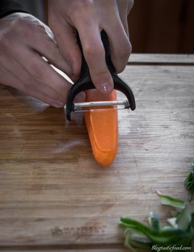 Someone using a speed peeler to peel a carrot into long thin strips.