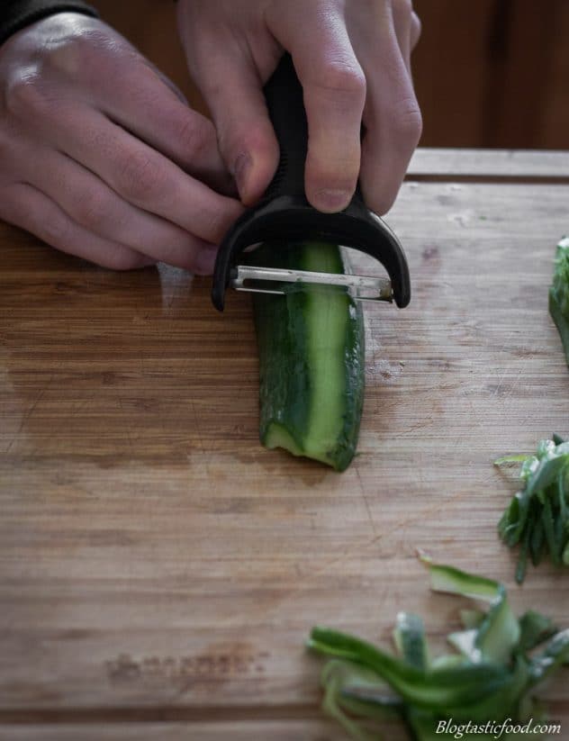 A cucumber being peeled into long strips on a chopping board.