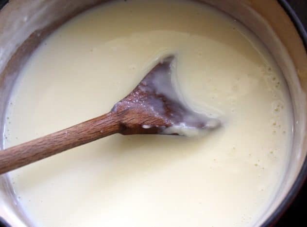 A close up photo of bechamel sauce in a pot with a wooden spoon stirring it.