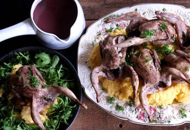 An overhead photo of roasted quails on a bed of polenta with a pot of gravy on the side. 