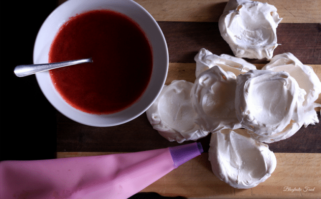 An overhead photo of mini meringues, a piping bag and a bowl of strawberry coulis on a wooden board.