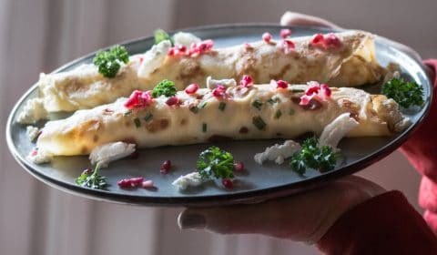 Spinach and ricotta filled crepes served on a fancy plate, garnished with pomegranate seeds, parsley and goats cheese.