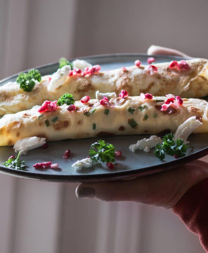 Spinach and ricotta filled crepes served on a fancy plate, garnished with pomegranate seeds, parsley and goats cheese.
