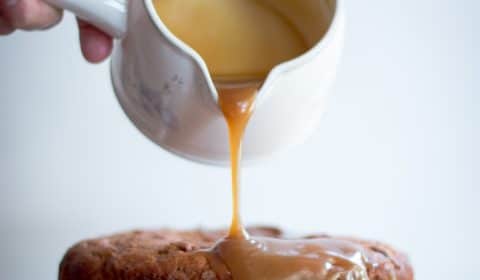 Someone pouring butterscotch sauce onto a sticky date pudding on a cake stand.