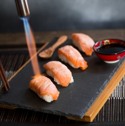 Someone using a blowtorch to sear the salmon sushi nigiri on a platter with a mini bowl of soy sauce.