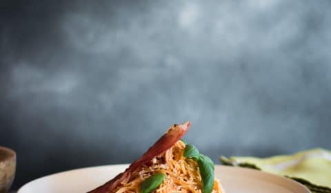 An eye level shot of angel hair napolitana pasta garnished with basil and a piece of crispy prosciutto.