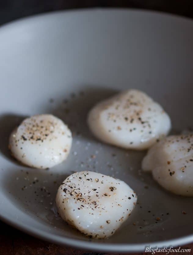 Raw scallops on a plate that have been seasoned with salt and pepper. 