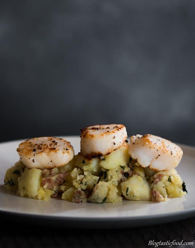 Seared scallops on top of crushed potatoes served on a white plate. 