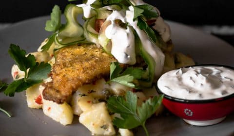 A photo of polenta coated fish on a bed of dressed potatoes garnished with cucumber and sour cream sauce.