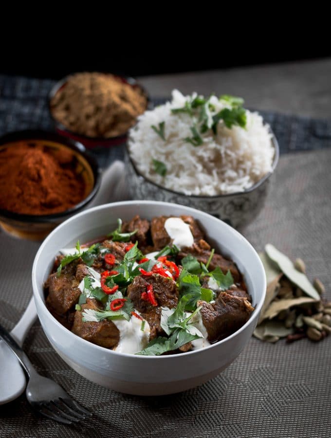 A bowl of Lamb curry with rice and spices in the background.