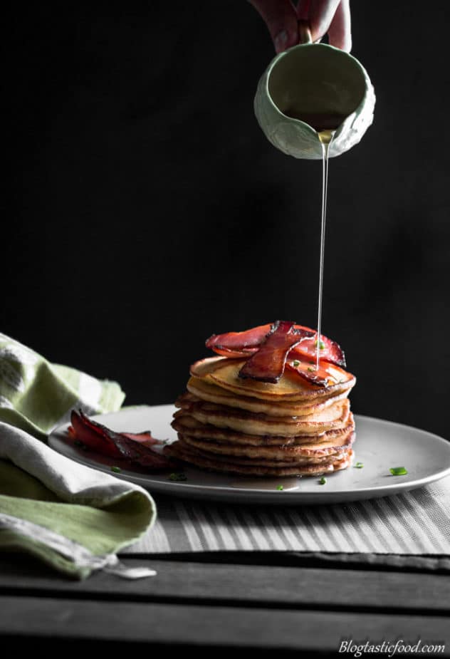 A stack of thick, fluffy pancakes with crispy bacon and maple syrup being poured over the top.