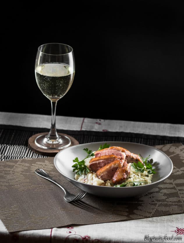 A bowl of herb risotto with pan seared duck breast and a glass of white wine in the background. 