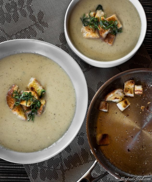 A overhead photo of cauliflower and leek soup in 2 bowls with a pan of croutons. 