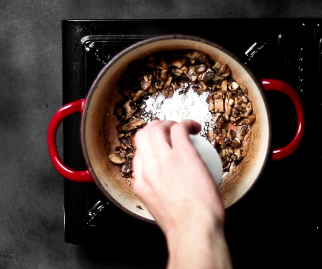 A photo of someone adding flout to a pot filled with sauteing mushrooms.