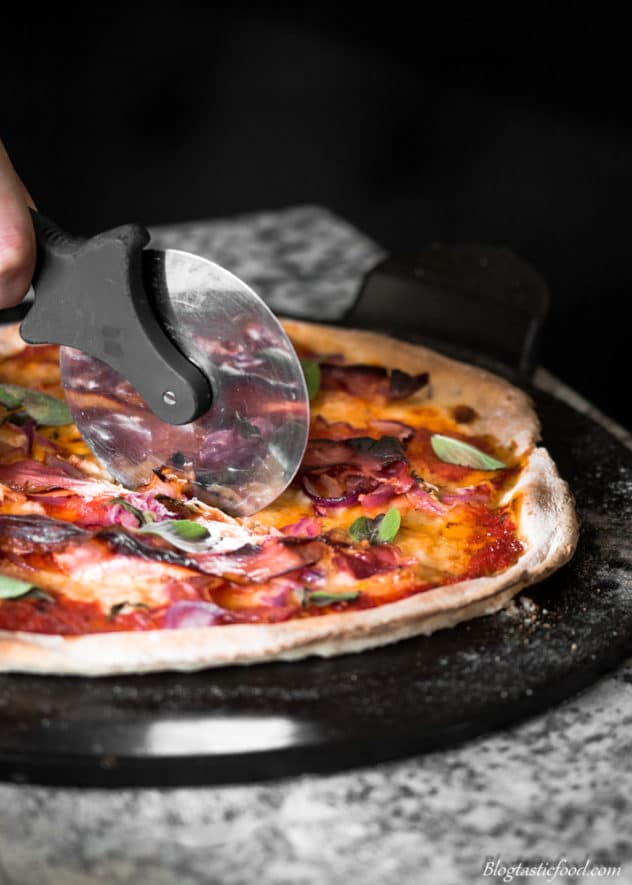 A photo of crispy prosciutto Margherita pizza being sliced with a pizza cutter.