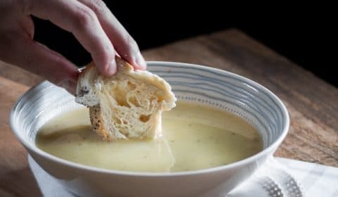 A bowl of creamy vegan potato soup with a piece of bread being dunked into it.