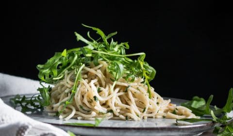 An eye level photo of aglio e olio pasta with rocket leaves on top. Served on a nice, flat plat on a white piece of a cloth.