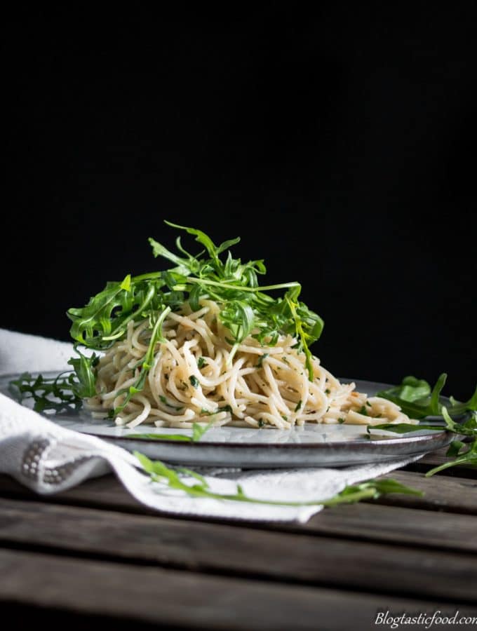 An eye level photo of aglio e olio pasta with rocket leaves on top. Served on a nice, flat plat on a white piece of a cloth.