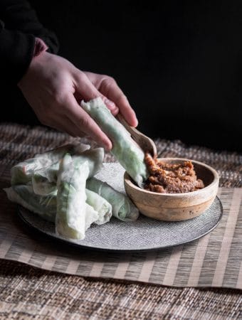 A dark, moody photo of fresh Vietnamese spring rolls on a plate and a small wooden bowl filled with peanut satay sauce. In the photo someone is also spooning some peanut satay sauce on one of the spring rolls.