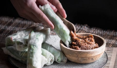 A dark, moody photo of fresh Vietnamese spring rolls on a plate and a small wooden bowl filled with peanut satay sauce. In the photo someone is also spooning some peanut satay sauce on one of the spring rolls.