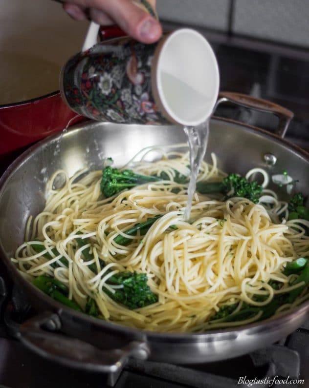 Someone pouring in some pasta water in a pan filled with spaghetti and broccolini.