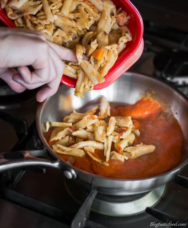 A photo of cooked pasta being added to a pan of red pepper and tomato sauce. 