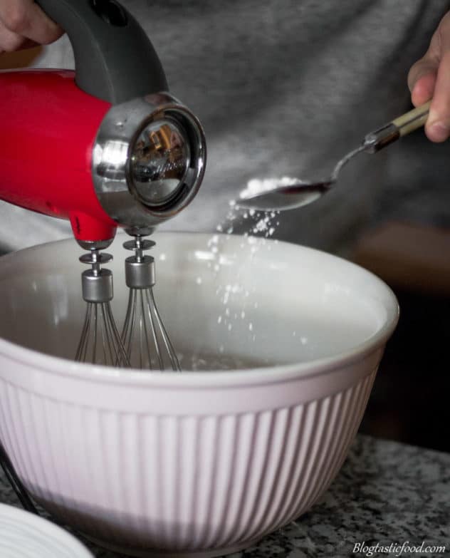A photo of someone spooning icing sugar in a bowl of meringue as it's being whisked.