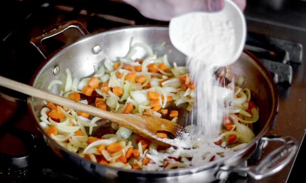 A photo of flour being added carrots and onion frying off in a pan. 