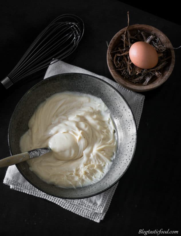 An overhead photo of a bowl of mayonnaise, a whisk and an egg in a small wooden bowl.