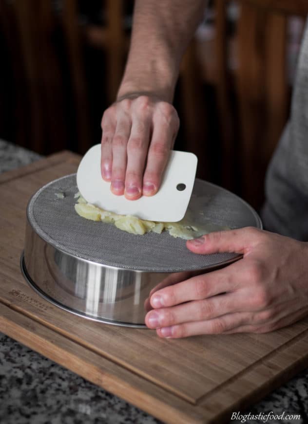 A photo of someone pressing boiled potatoes through a drum sieve.