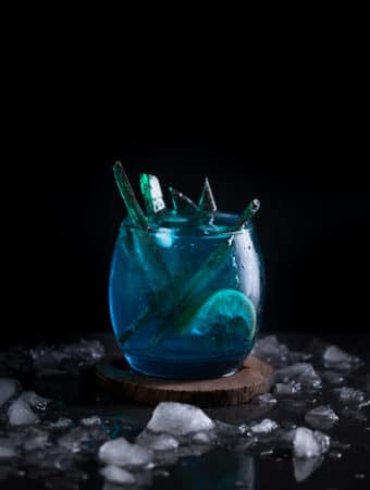 A photo of a blue white walker themed cocktail with green candy inside of it, served on a wooden coaster.