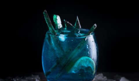 A photo of a blue white walker themed cocktail with green candy inside of it, served on a wooden coaster.
