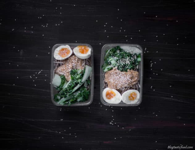 An photo of 2 plasitc containers filled with cooked soba noodles, boiled eggs, sauteed bok choy, peaut sauce and sesame seeds.