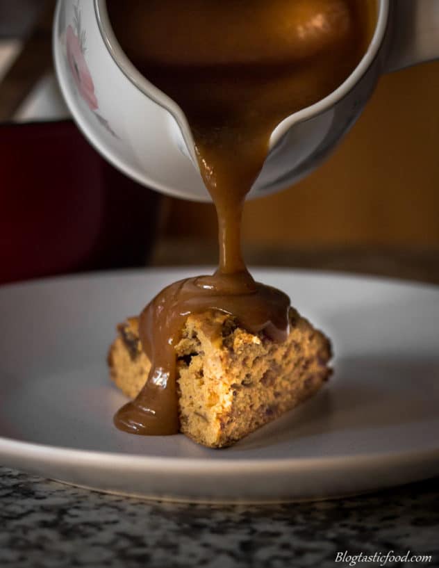 A portion of sticky date pudding on a white plate, with butterscotch sauce being poured on top.