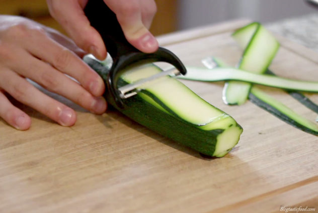 Someone using a speed peeler to peel thin slithers out of zicchini.