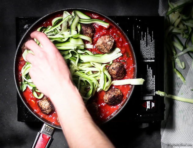 Someone adding zoodle to non stick pan filled with meatballs and tomato sauce.