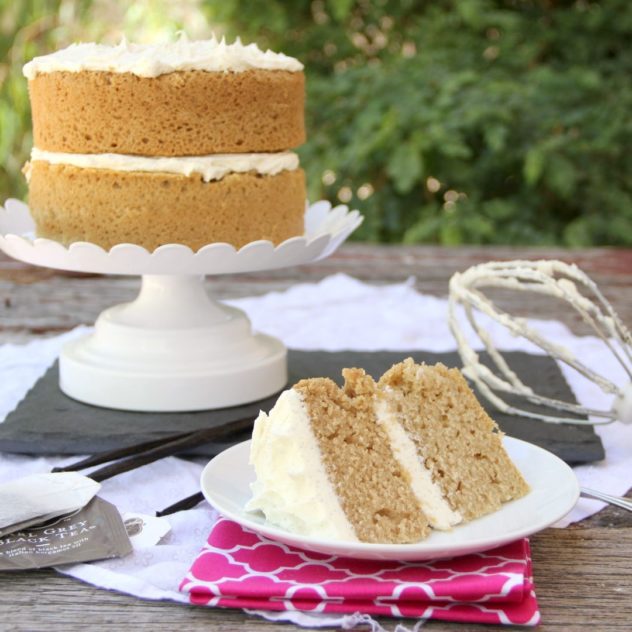 A portion of earl grey cake with vanilla frosting on a white plate, with the rest of the cake on a stand in the background. 