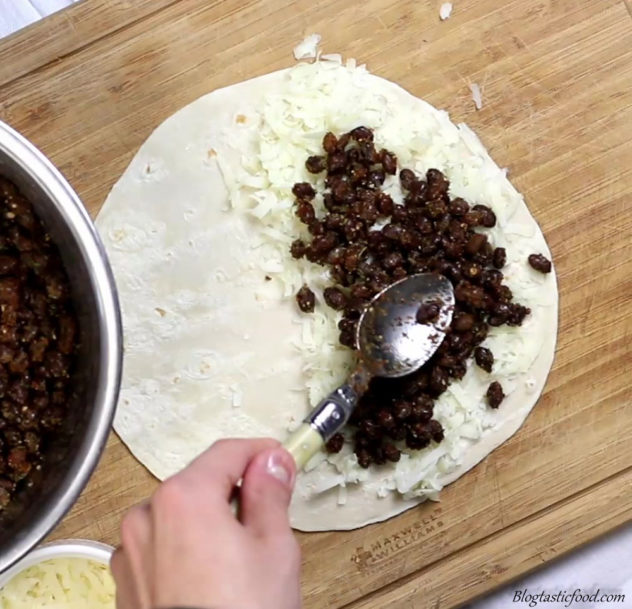 a photo of someone placing spiced beans over a layer of cheese over only half of the tortilla.