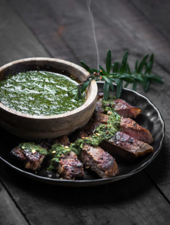 Slices of sirloin steak on a black plate beside a bowl of chimichurri sauce.