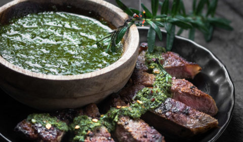 Slices of sirloin steak on a black plate beside a bowl of chimichurri sauce.