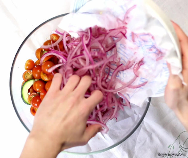 Marinated red onions being added to a bowl filled with cherry tomatoes and sliced cucumbers.