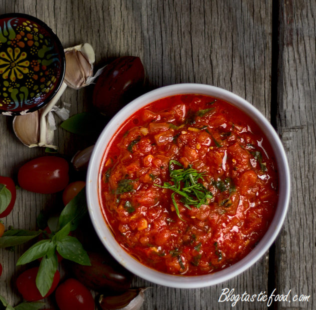 A white bowl filled with tomato sauce, beside cherry tomatoes, basil, garlic and a mini bowl of olive oil.